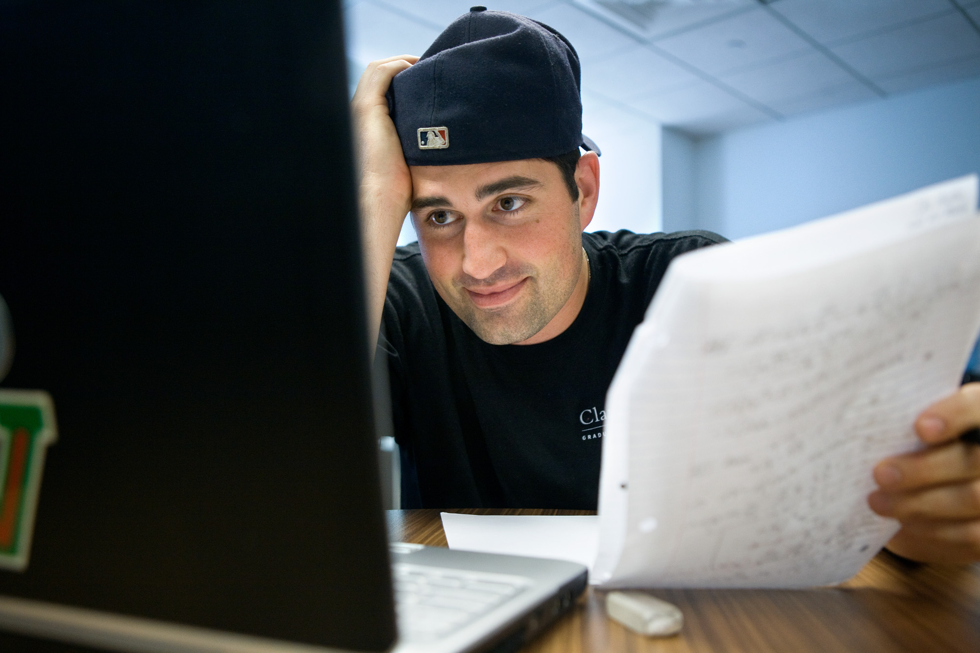 an operations research student wearing a backwards MLB cap holds a piece of paper while observing a laptop screen