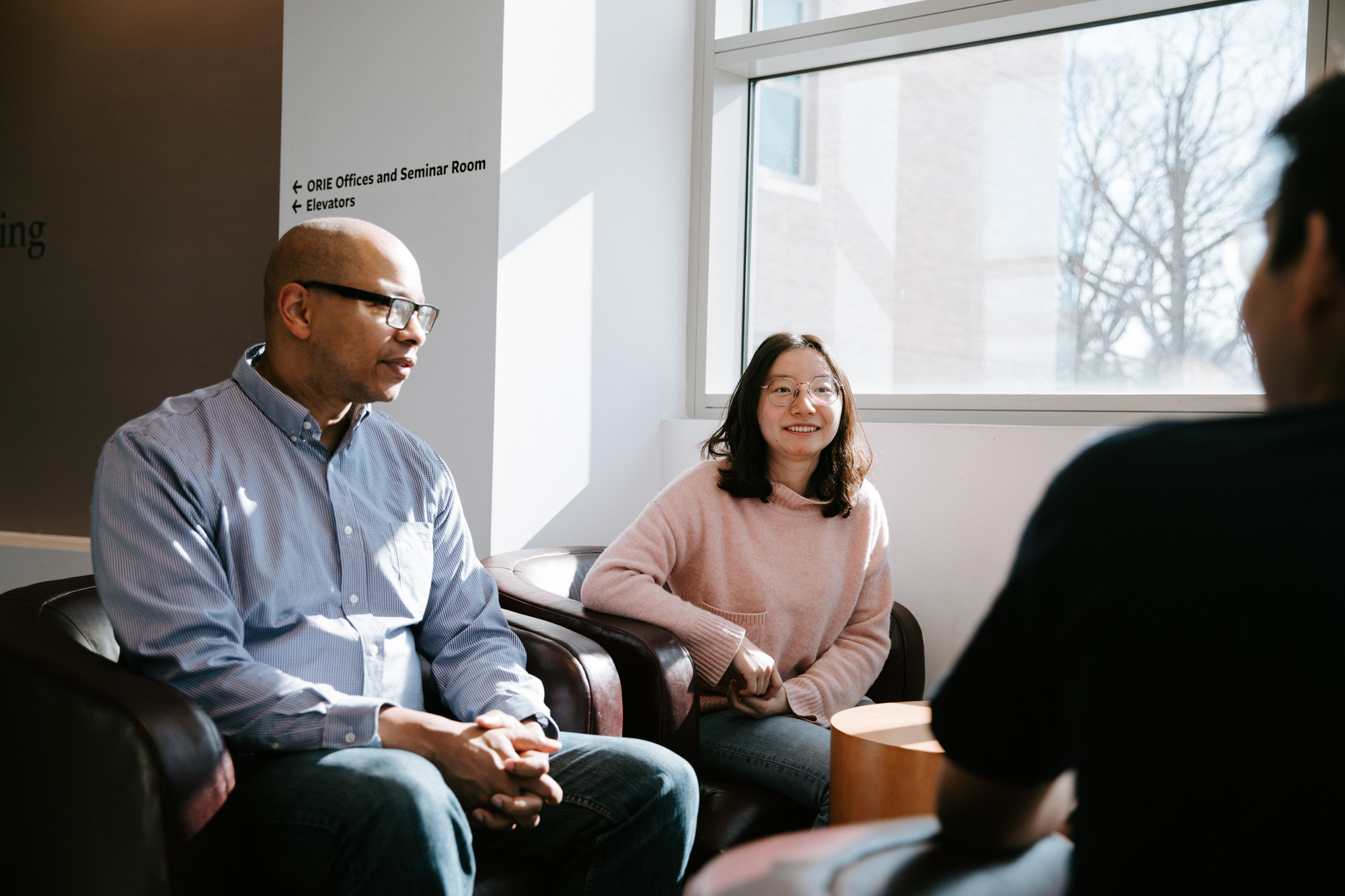 ORIE Professor Mark Lewis seated in Rhodes Hall as two seated students talk.