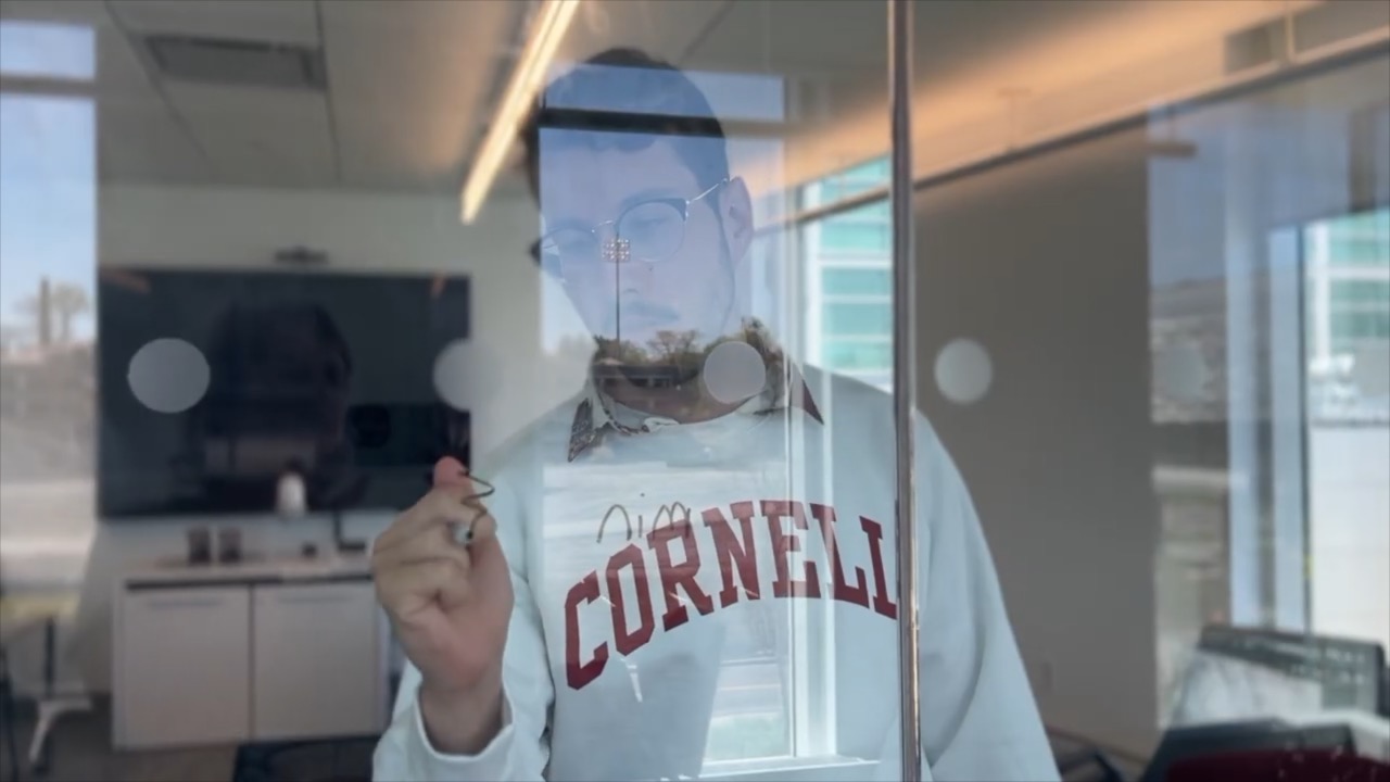 an operations research student wearing a Cornell sweatshirt writes onto a glass wall