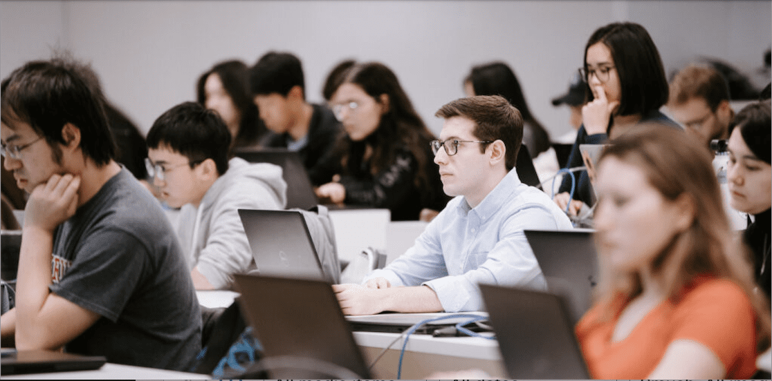 Rows of students sit in a lecture hall with laptops open in front of them.
