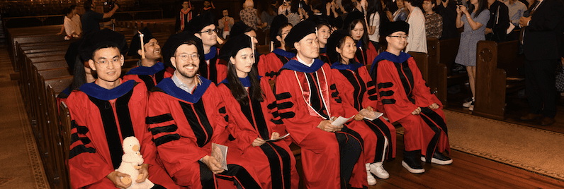 Smiling students in red graduation robes and black caps sit in Sage Chapel.