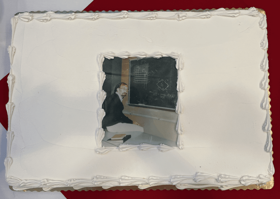 A cake with white frosting. In the center of the cake is an edible portrait of Fulkerson teaching by a blackboard.