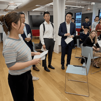 A woman stands with papers in her hands. Other people stand lloking at her.