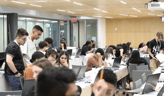 Students in a classroom work in groups or alone at laptops. Some are sitting, some are standing.