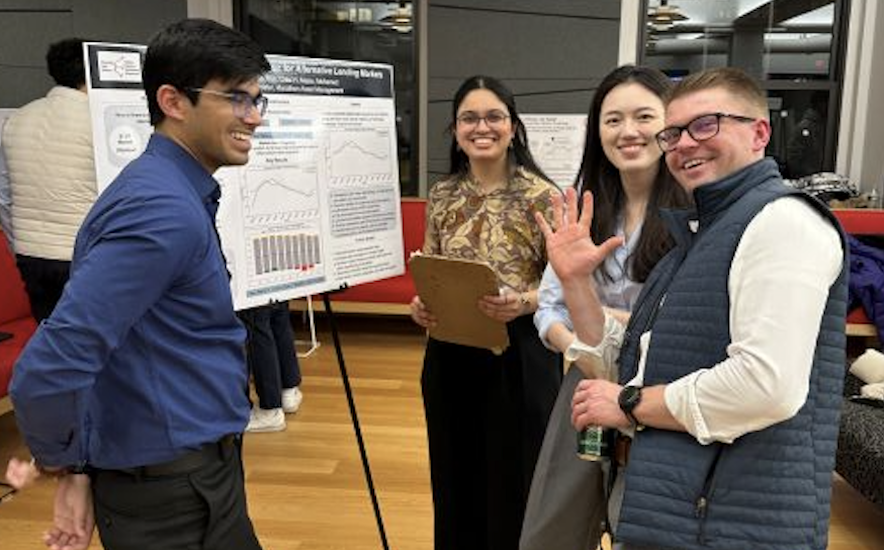 4 people stand in front of a poster on an easel and smile while looking at the viewer.