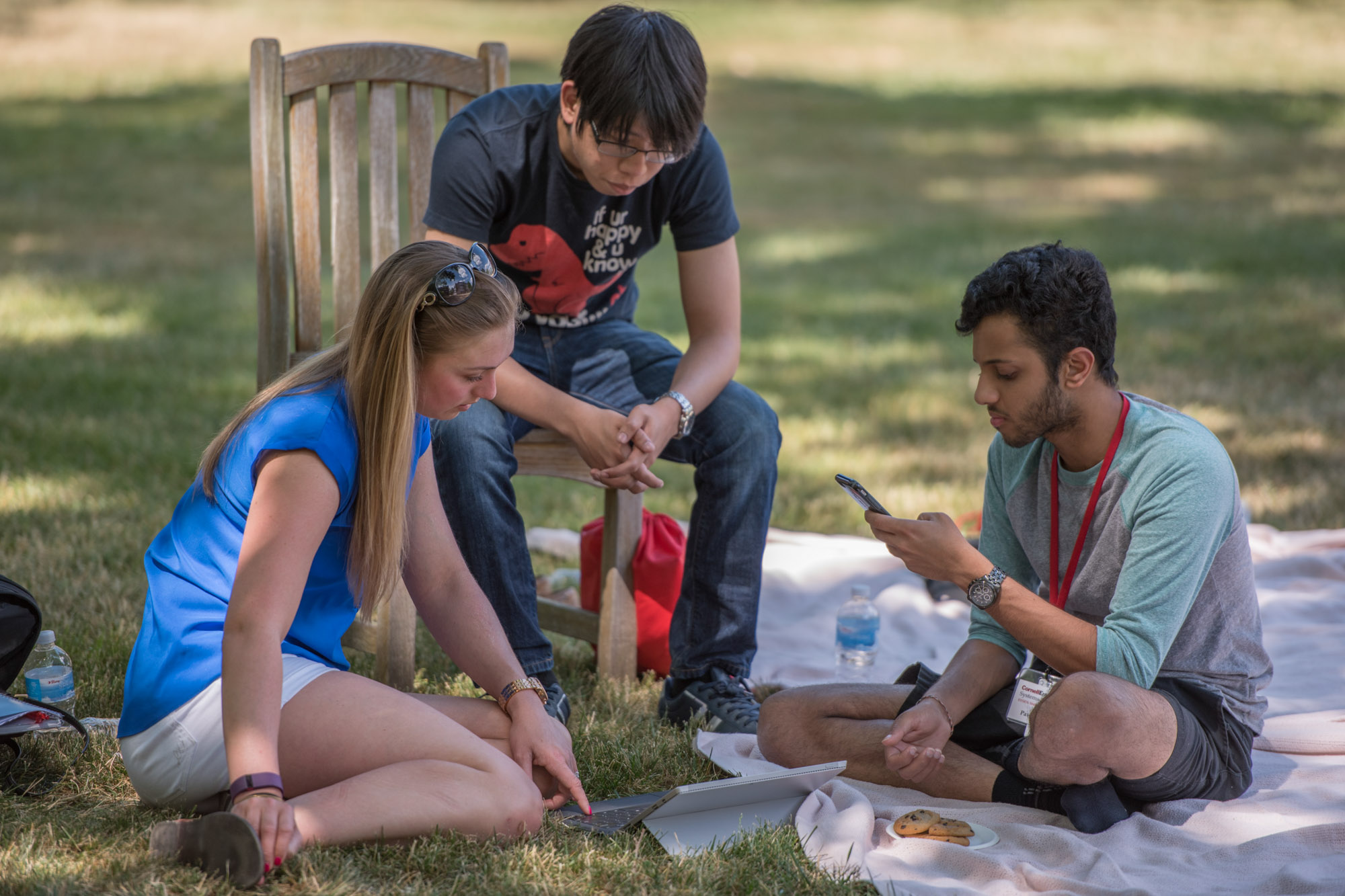 Two SYSEN 5940 students sit on the ground while a third sits in a chair, enjoying the summer weather outside on the Cornell campus