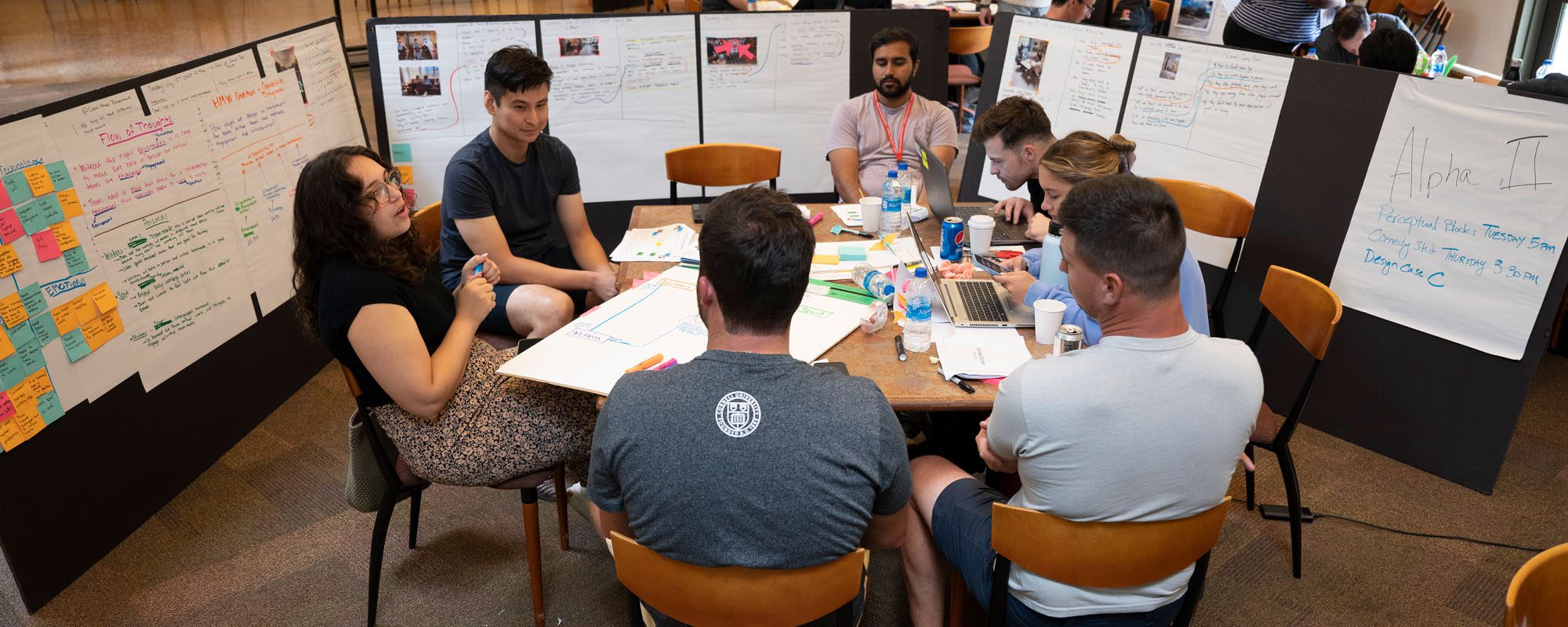 six students (men and women) work around a table. Surrounding them are easels filled with posters and sticky notes.