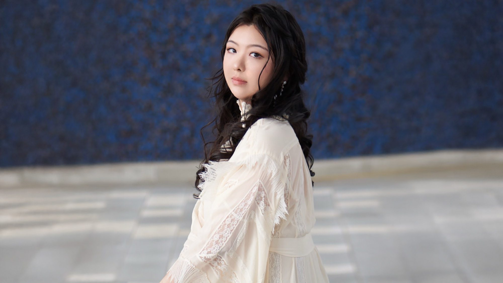 woman sitting against a blue wall, with head turned over shoulder to look at camera