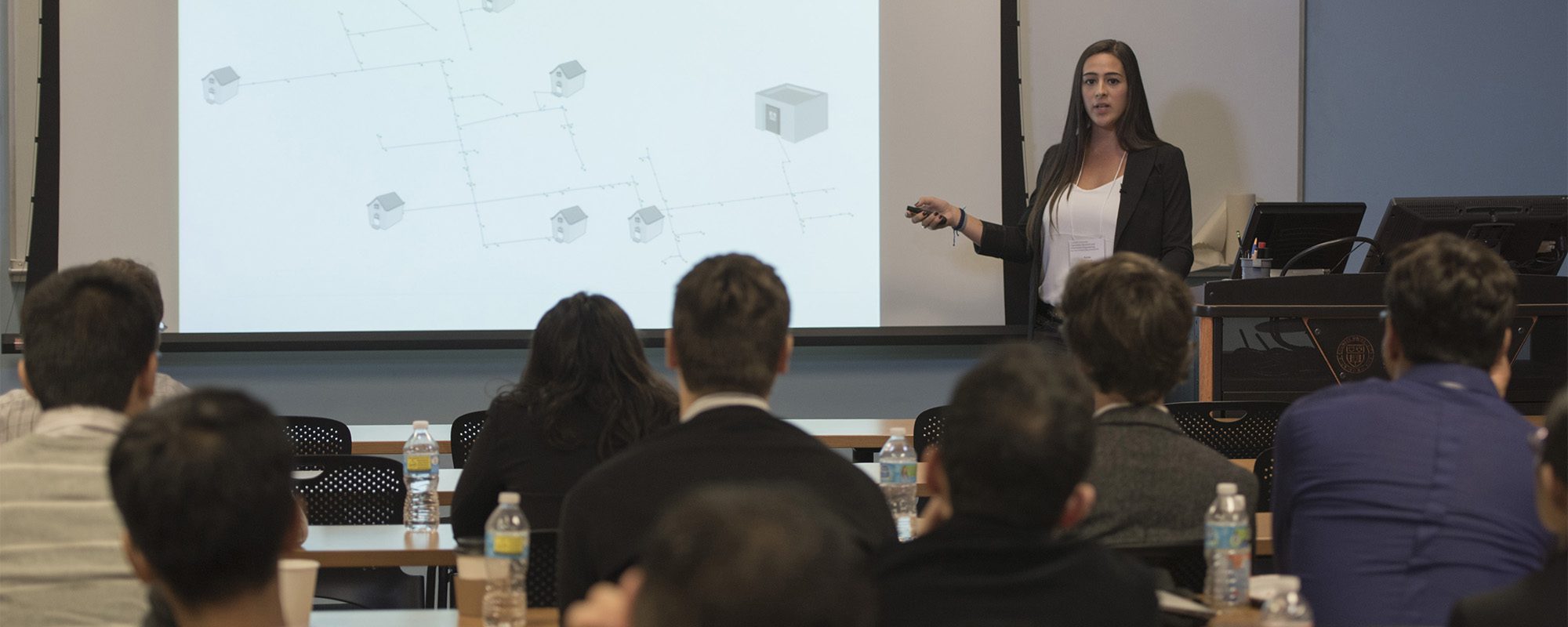 A woman stands in front of a room of people pointing towards a slide that is displayed on the screen.