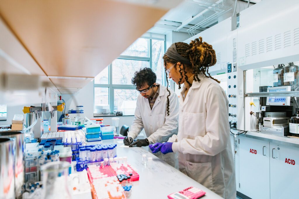 Two researchers in lab coats and gloves work together surrounded by test tubes and other equipment