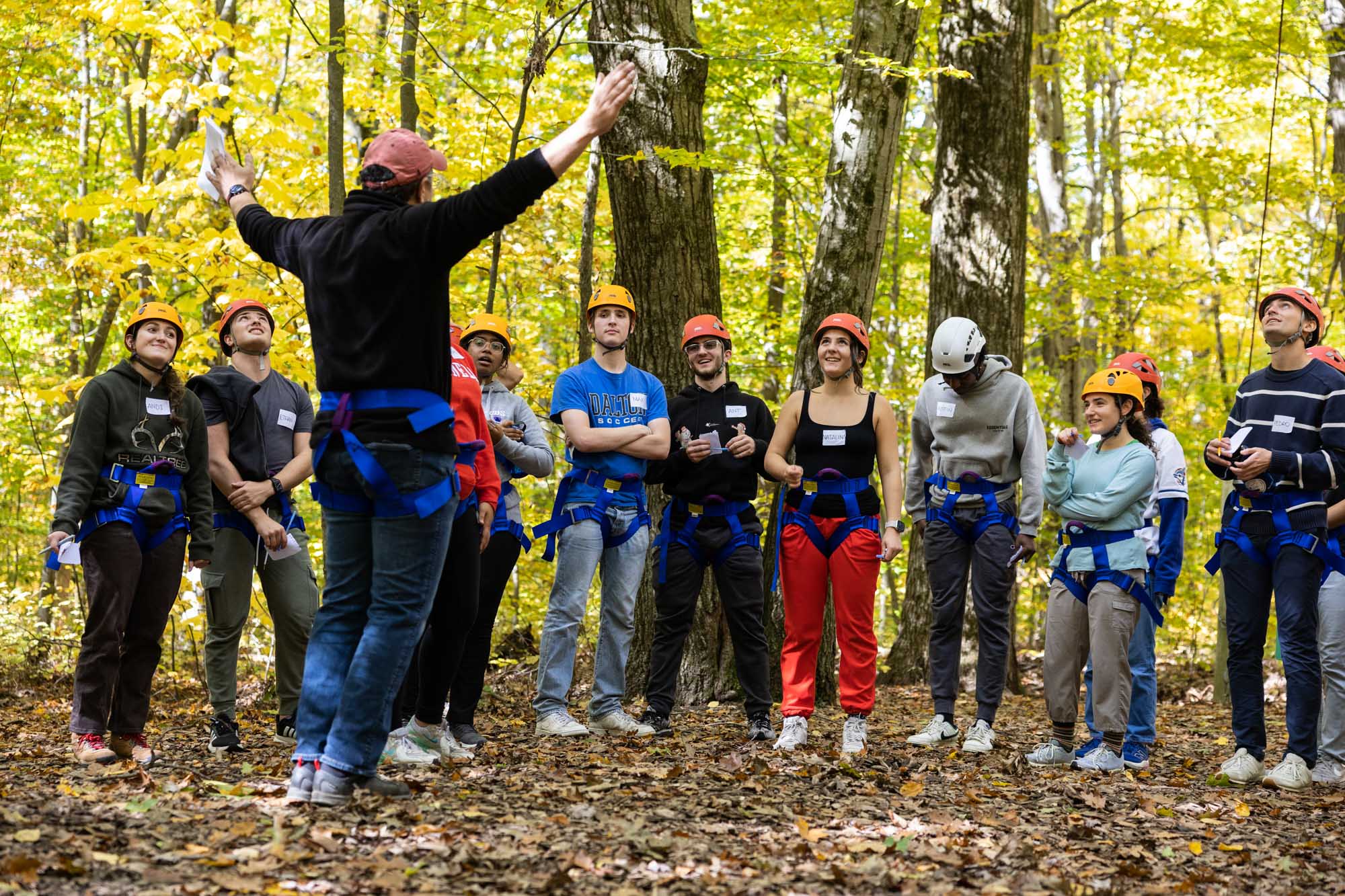 Instructor raises arms in a Y shape as they speak to group of students wearing helmets and harnesses in the woods