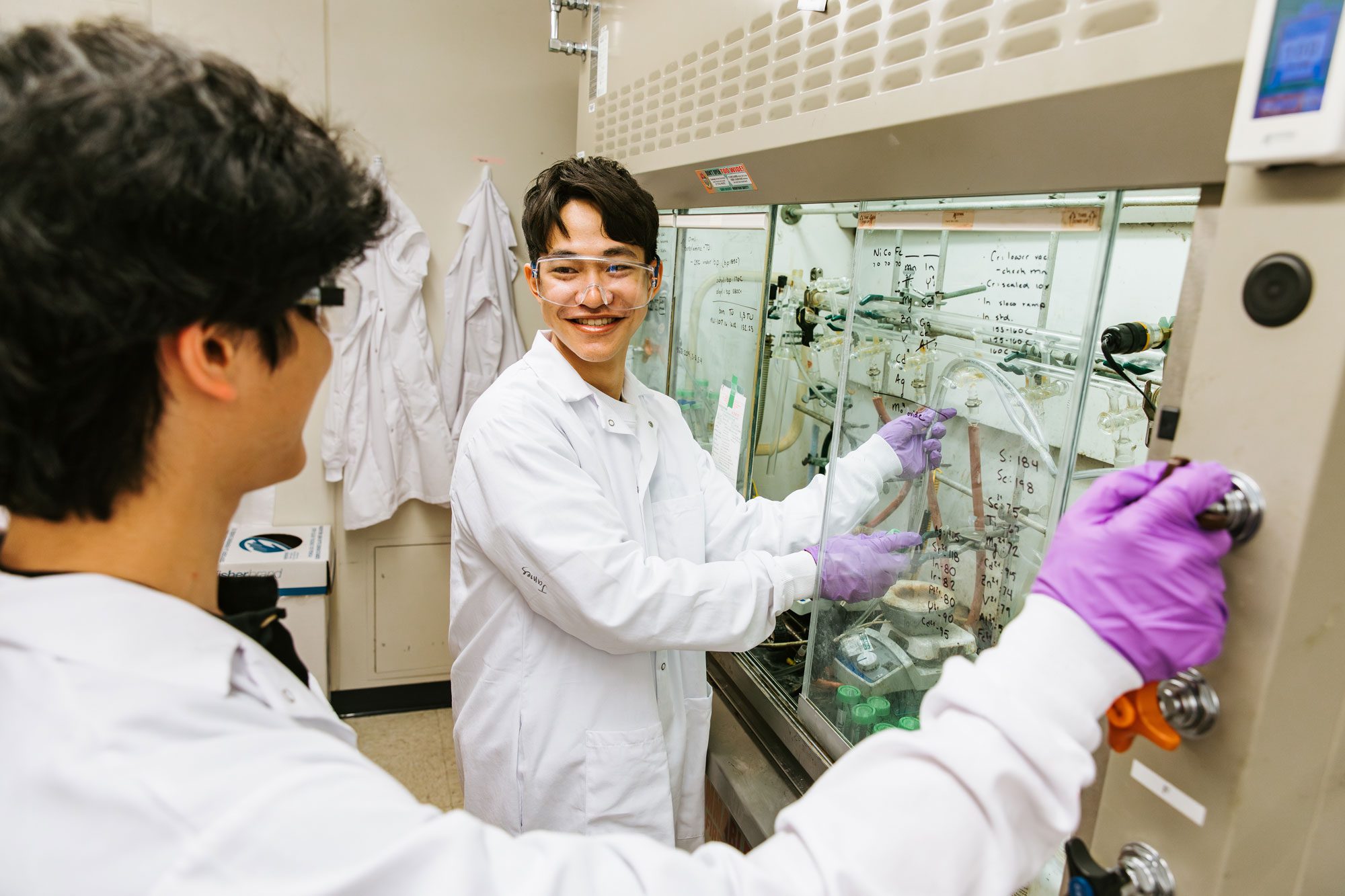Two students in protective clothing work with equipment behind glass case