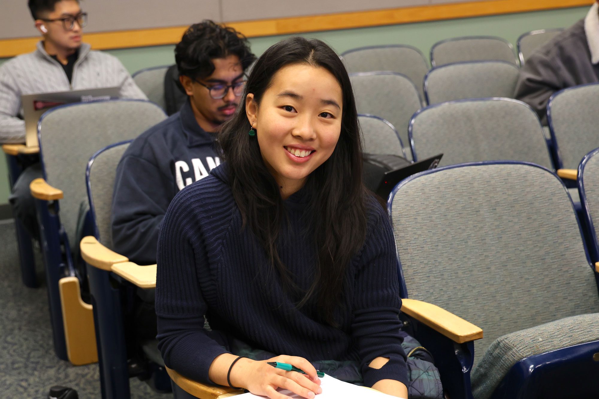 Student with notebook and pencil smiles, sitting in classroom with others