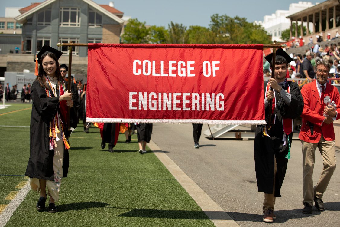Students in graduation caps and gowns carry Cornell College of Engineering banner as they walk on grass