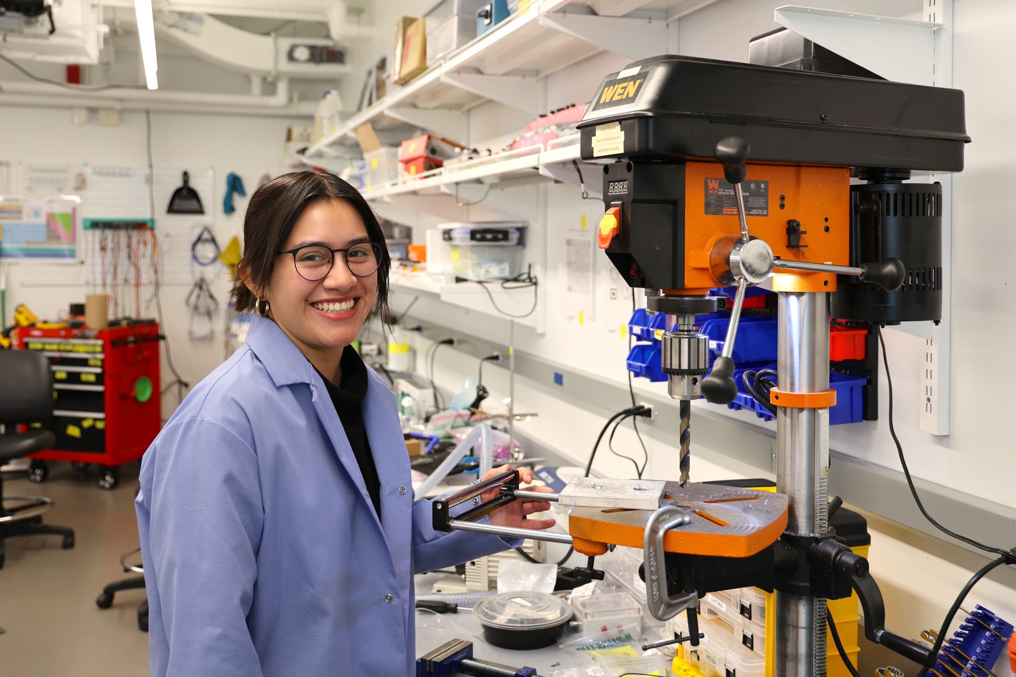 Student wearing lab coat smiles standing by machinery in lab