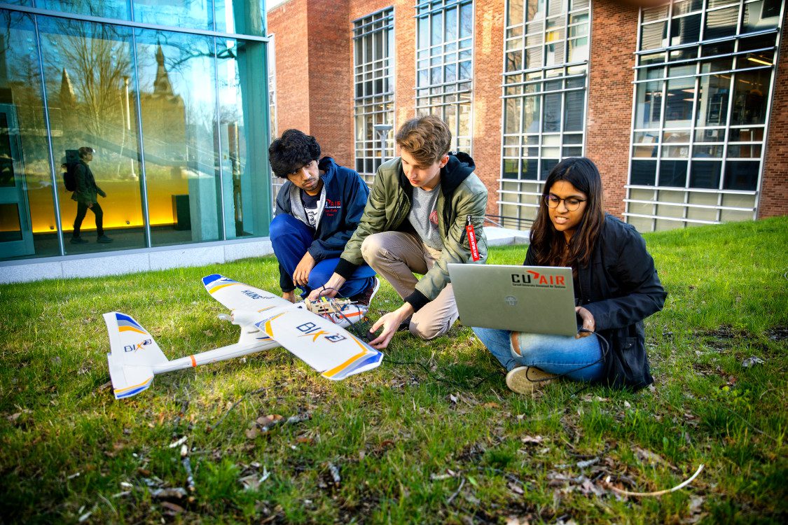 Three students sit on university lawn with small aircraft and laptop with sticker 