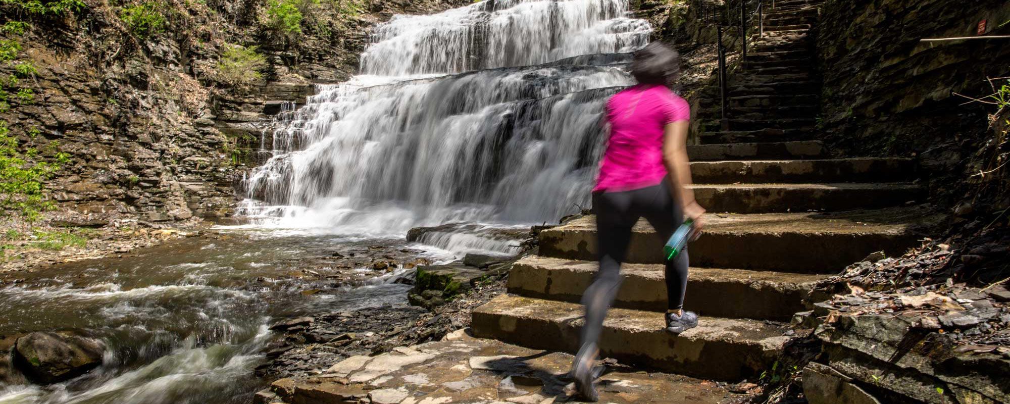 Hiker in a pink shirt climbing stairs with a waterfall in the background