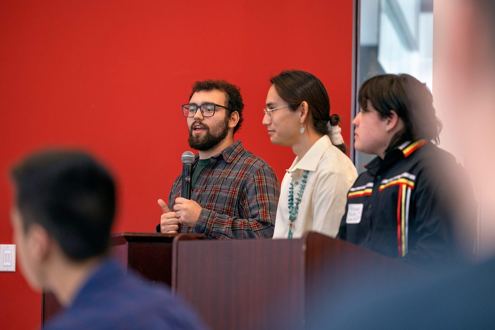 Male student speaking at event in a group of 3 in front of a red wall