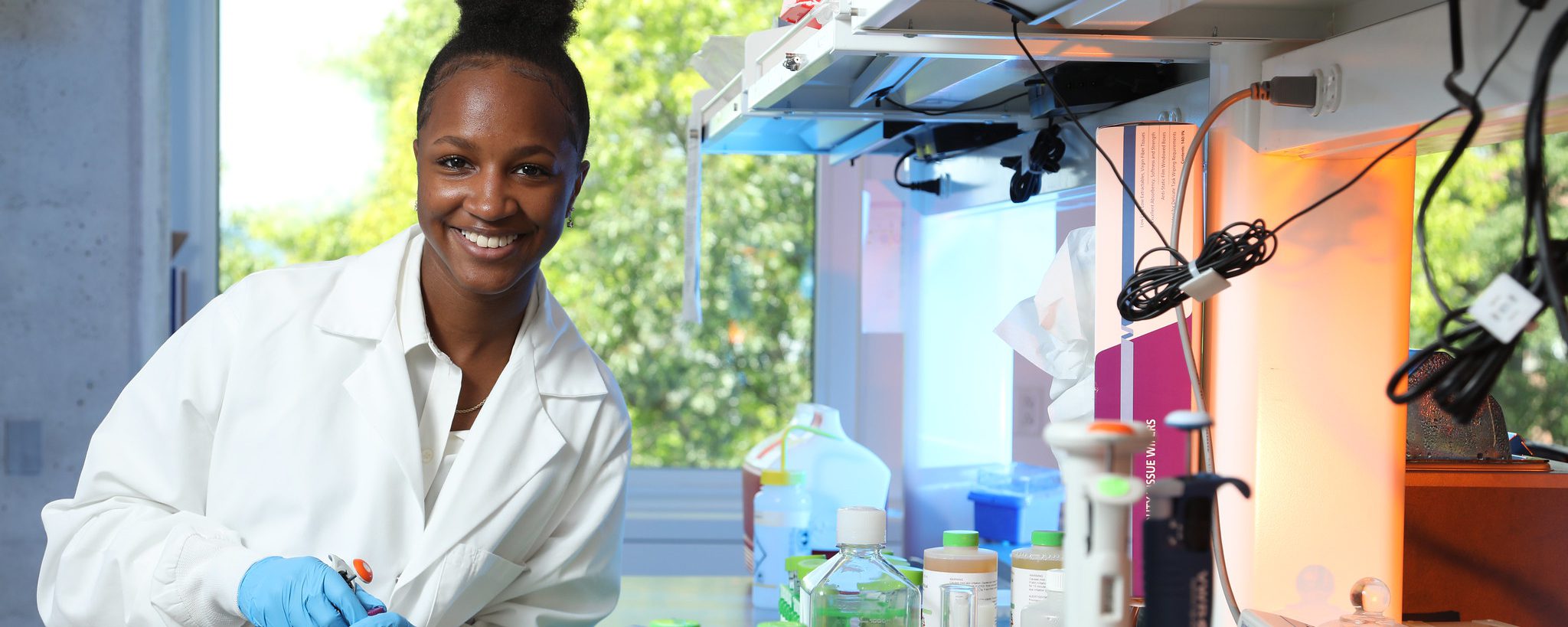 A student in a lab coat smiles for a photo in the lab while holding a pipette