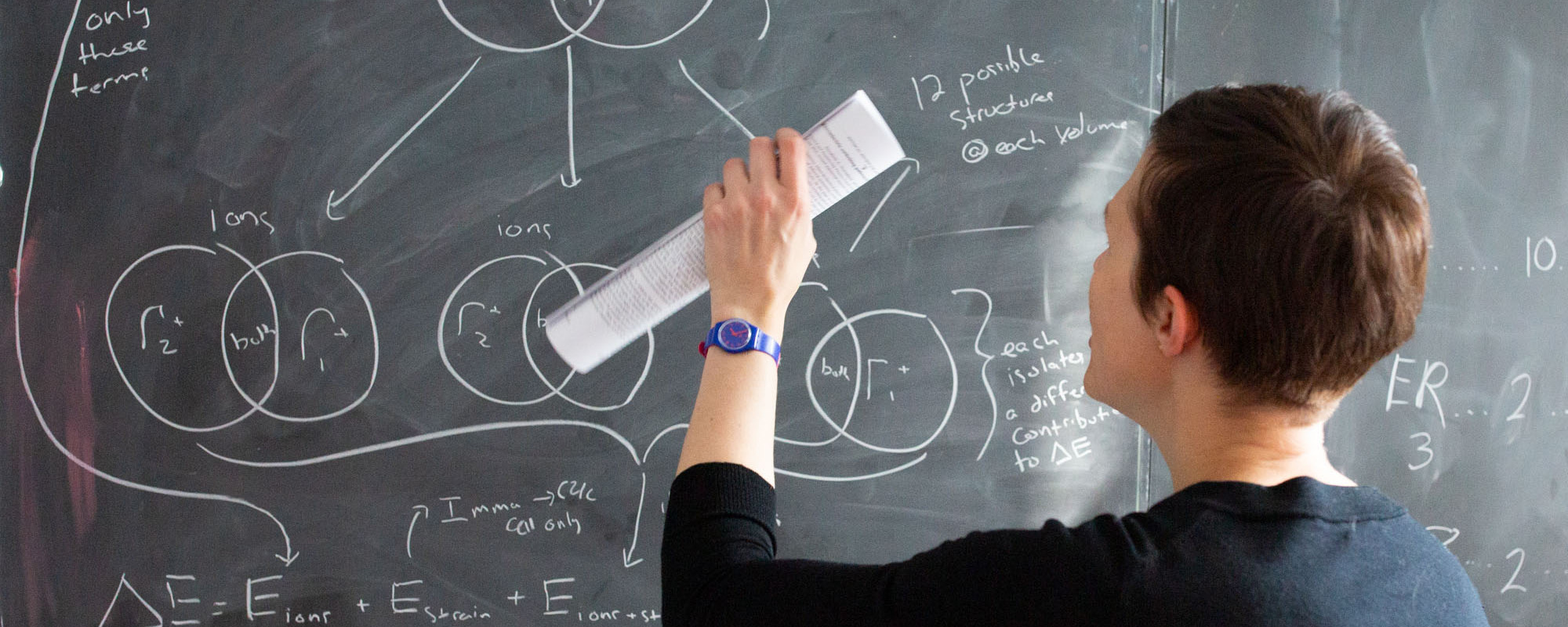 Female Professor in a black sweater holding a paper in front of a blackboard