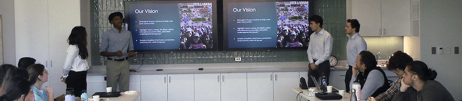 four students present on two screens to a conference room full of people