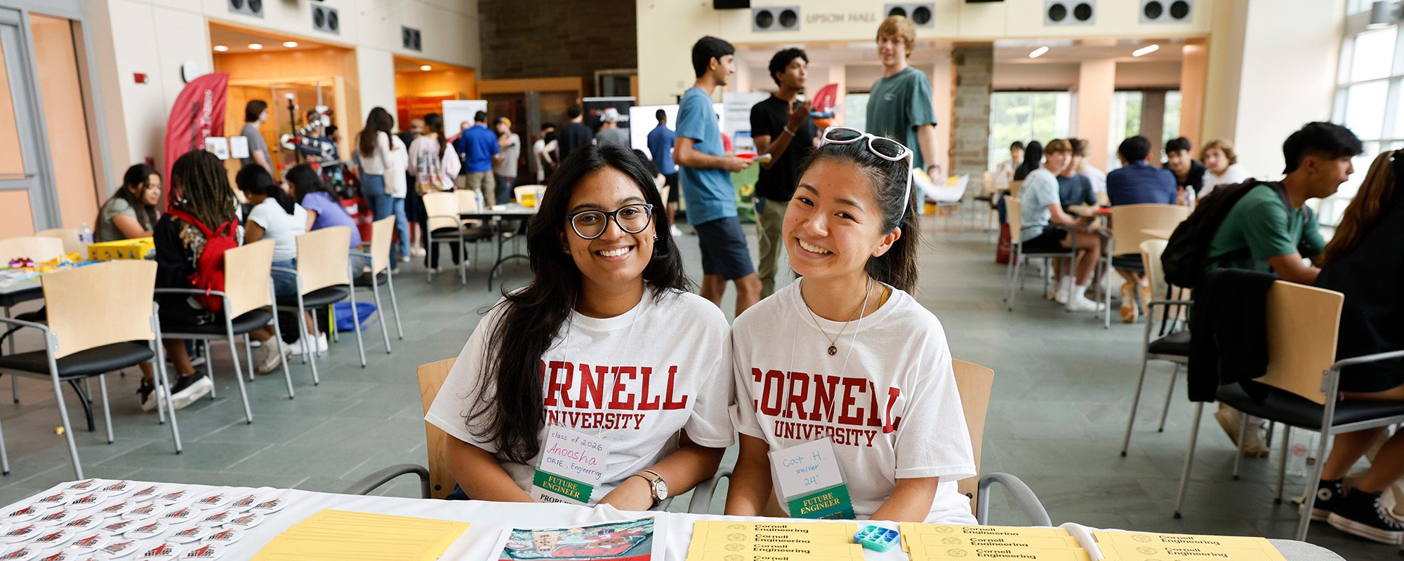 Two students wearing Cornell University shirts sit a table to welcome new students in Duffield Atrium