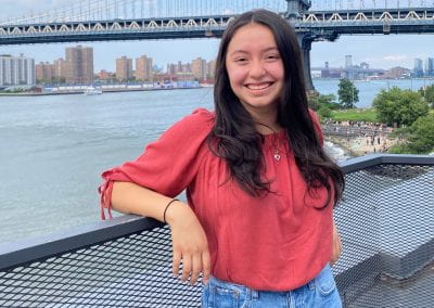 Student smiles, leaning against river railing with bridge in background