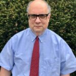 Man in glasses, shirt and red tie outside in front of greenery