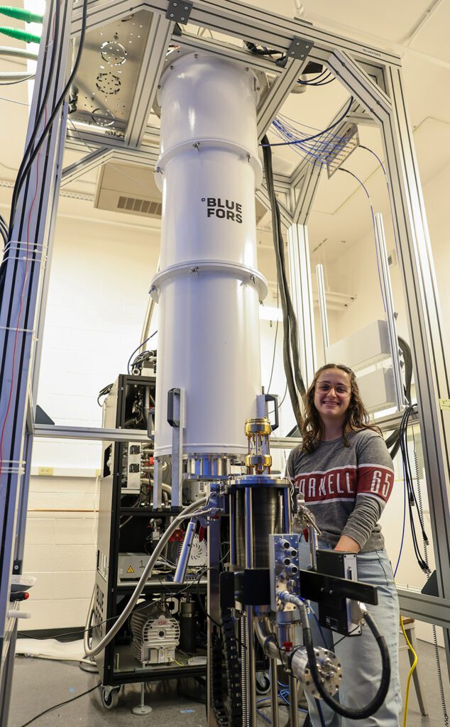 A woman stands next to a large white tube nearly twice her height, which houses a cryostat.