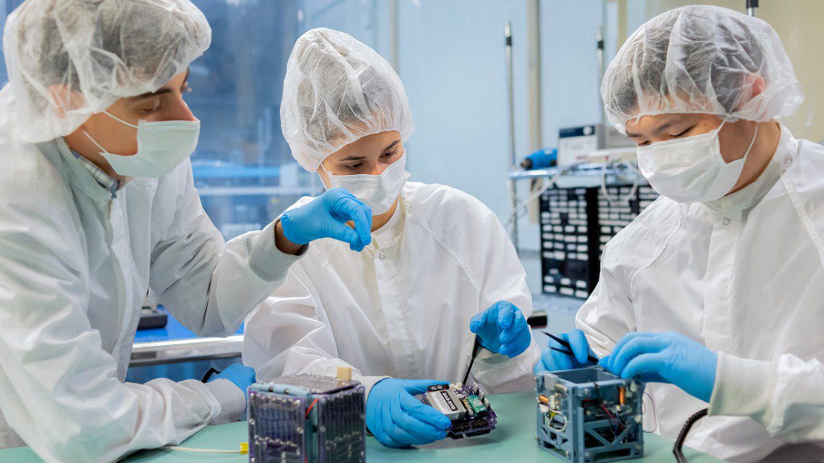 three students in a cleanroom observe a small spacecraft