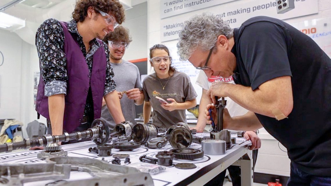 three students watch as a professor assembles part of an engine