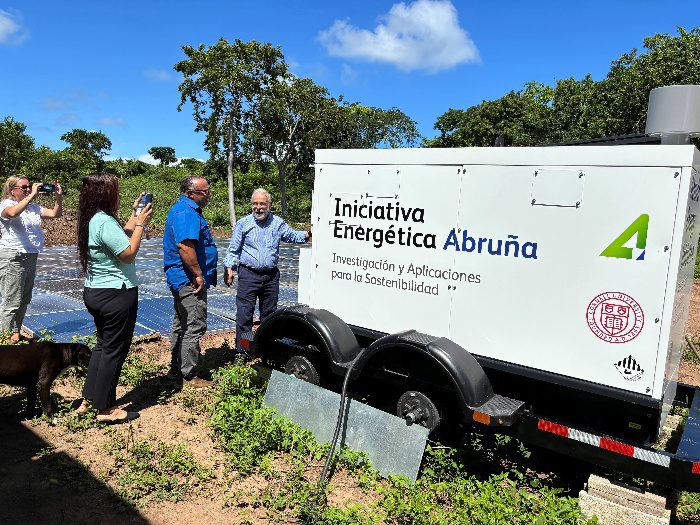 Four people stand to the left of a large whiite box on wheels. These words in Spanish are written on the side of the box: "Iniciativa Energética Abruña, Investigación y Aplicationes para la Sostenibilidad"