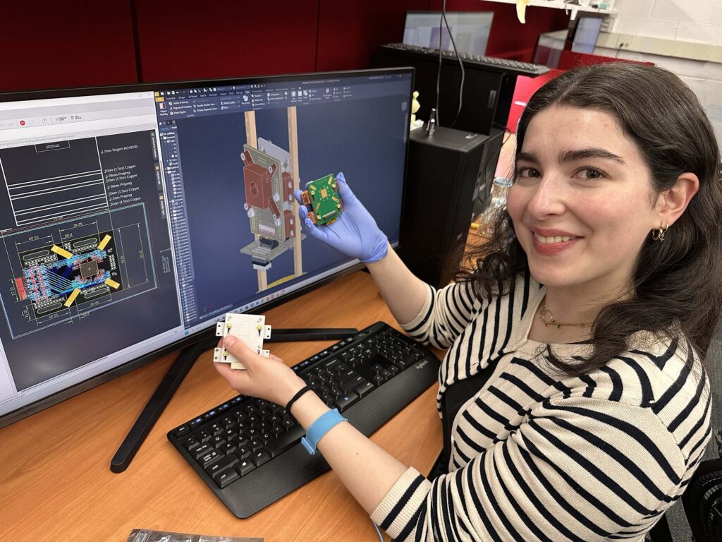 A woman sits in front of a computer in which she has generated CAD diagrams of two devices she is holding. 