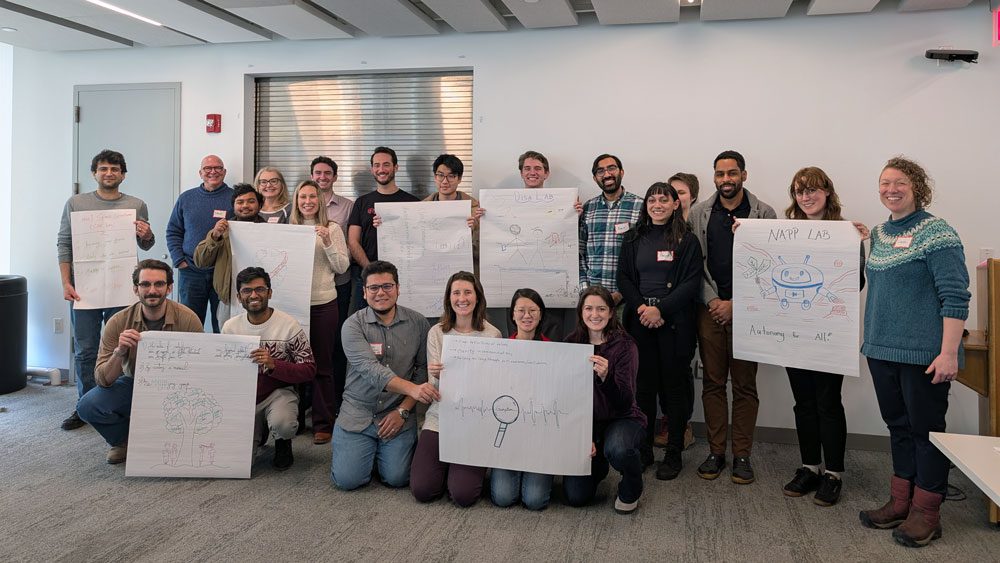 A group of twenty students and faculty stand in a group photo with severn students holding up large paper drawings representing labs