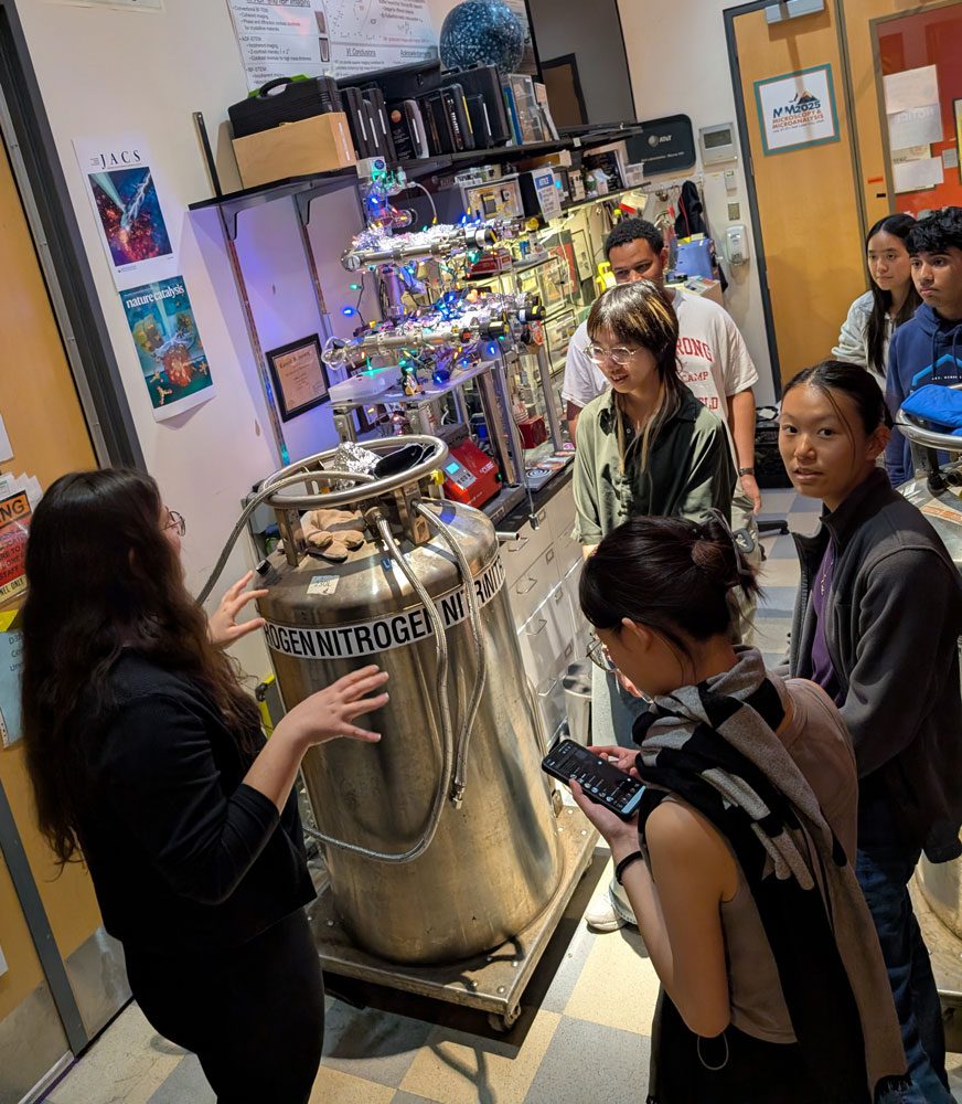 Students gather around metal canister with word "nitrogen" visible in classroom. Periodic table hangs from ceiling