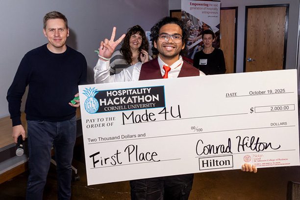 Smiling M.Eng. student Siddhant Dhende holds a giant check, makes a peace sign with his fingers, accepting a first place win at a Hospitality Hackathon event.