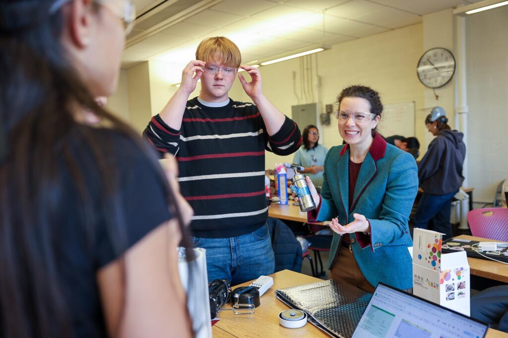 Allison Godwin smiling in classroom with her student adjusting goggles with student made devices