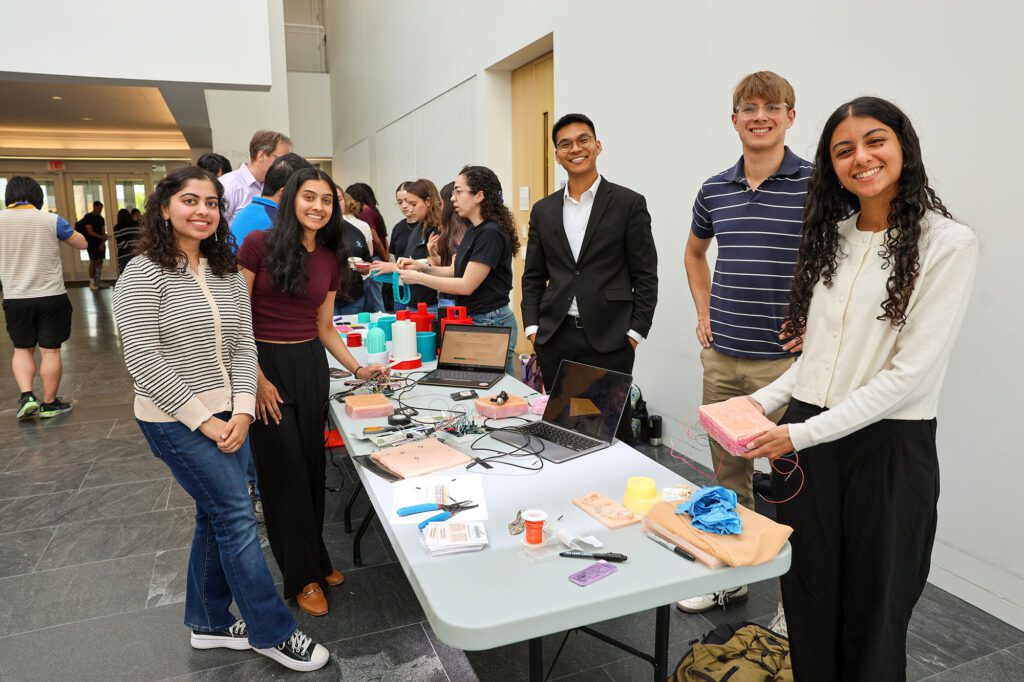 undergraduate biomedical engineering student marcus gamboa and his senior design team stand together with project in Weill Hall at the senior design showcase.