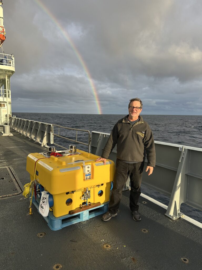 Geoff Abers stands on the deck of the R/V Thomas G Thompson, next to a yellow ocean-bottom seismometers that will be deployed on the sea floor around northwest New Zealand. There is a rainbow in the sky behind Abers.