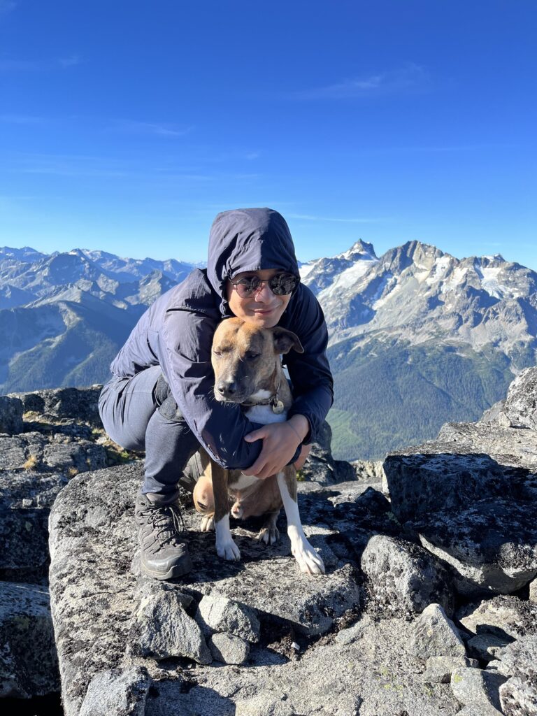 Tingan Li wears a wind jacket and hugs a dog high on a rocky mountain with a deep blue sky behind them.
